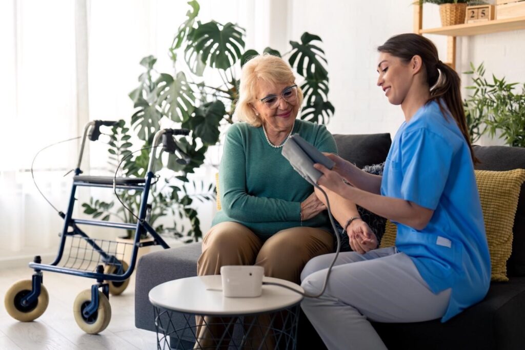 Female caregiver checking the blood pressure of a smiling elderly woman at home, with a walker in the background.