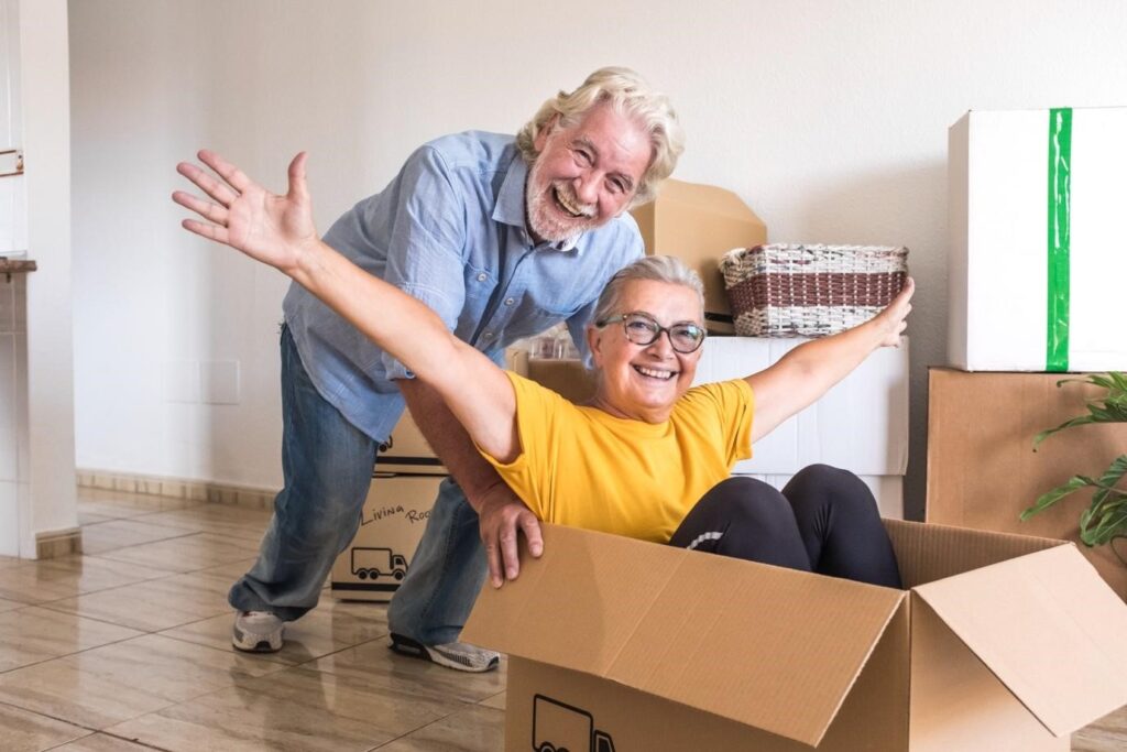 Cheerful senior woman sitting in a cardboard box with arms outstretched while her smiling partner pushes her playfully during a home move.