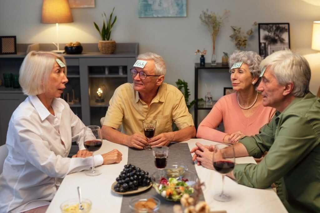 Group of cheerful seniors playing a word guessing game with sticky notes on their foreheads, enjoying wine and snacks around a table.