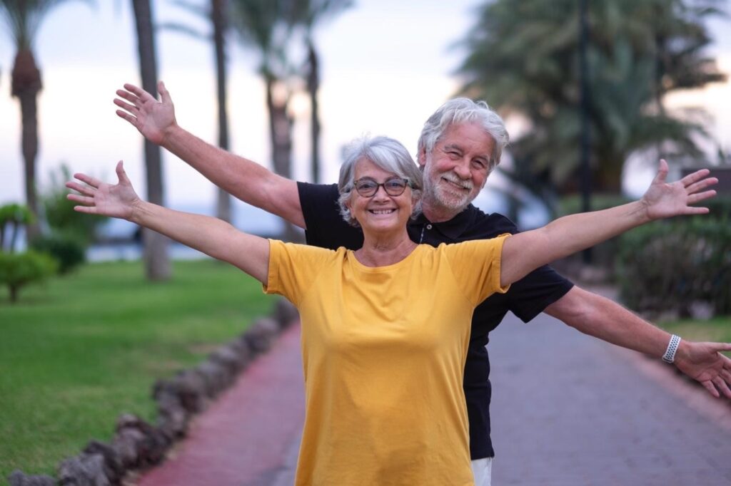 Happy senior couple smiling with outstretched arms while standing together on a scenic path lined with palm trees.