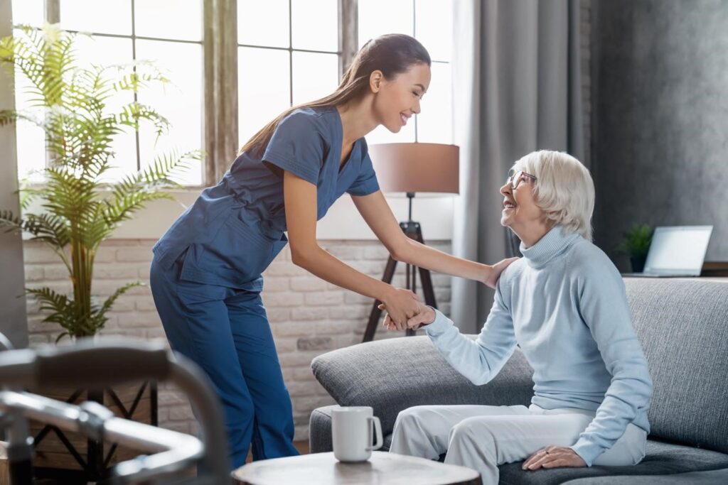 Young female caregiver warmly greeting an elderly woman sitting on a sofa in a cozy, well-lit living room.