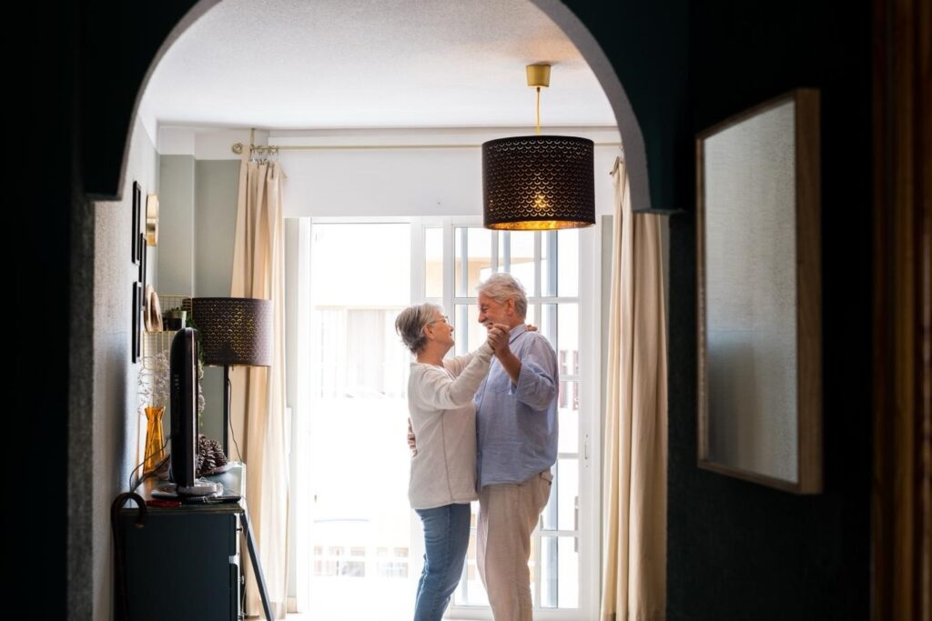 Happy elderly couple dancing together in a bright living room near a large window.