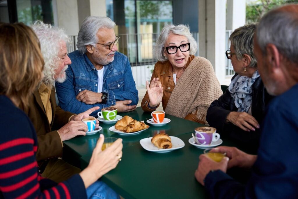 Group of senior friends enjoying coffee and pastries while having an animated conversation at an outdoor café table.