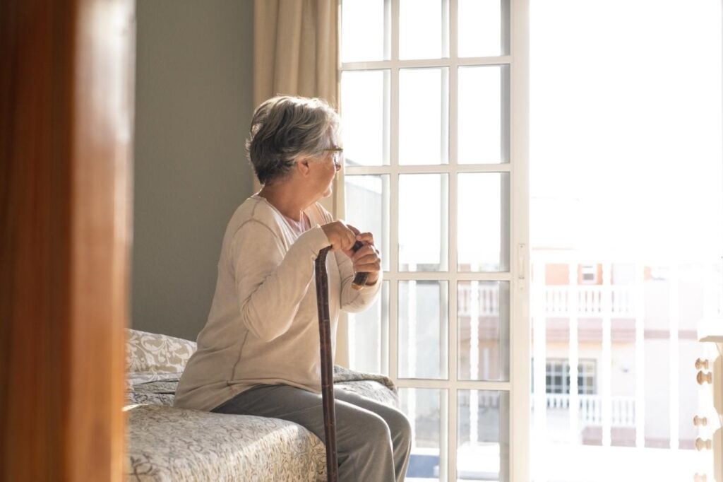 Elderly woman sitting alone on the edge of a bed, holding a walking cane and looking out through a bright window.