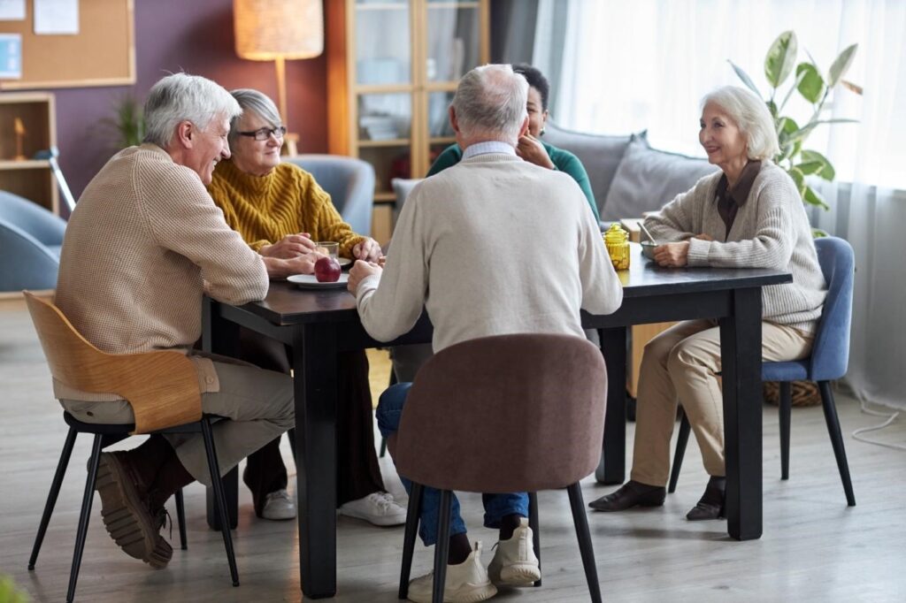 Group of elderly people sitting around a table in a cozy, well-lit room, engaged in cheerful conversation and enjoying each other’s company.
