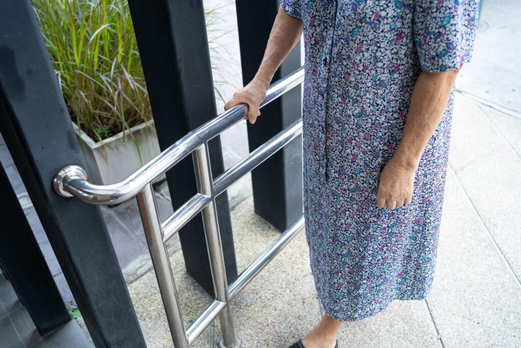 An elderly woman wearing a floral dress holds onto an outdoor stainless steel safety rail for support while walking.