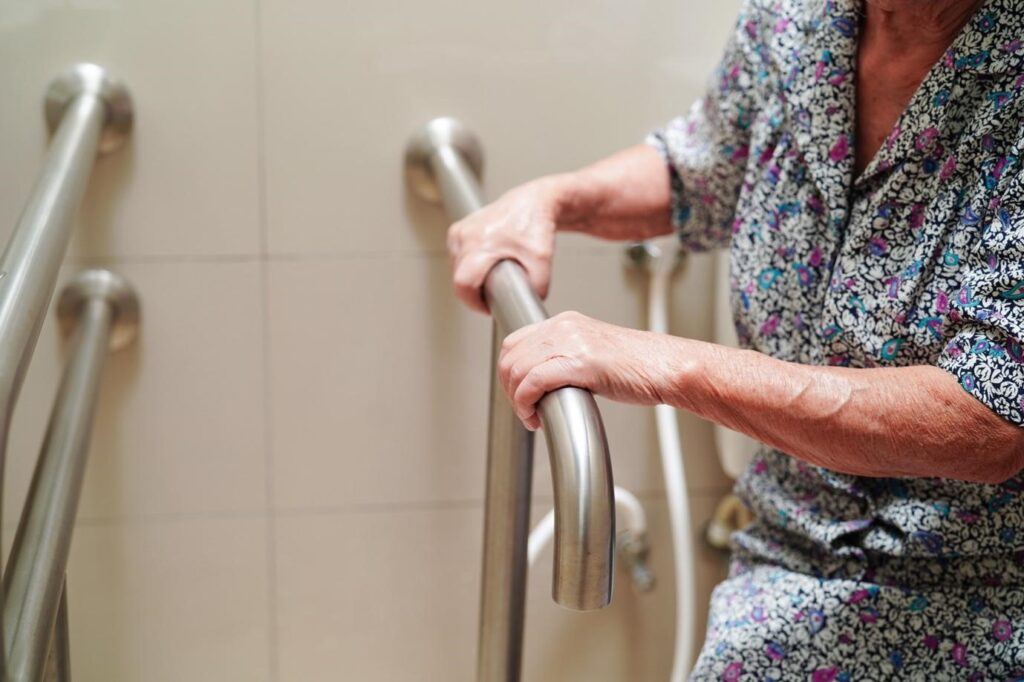 An elderly woman grips indoor bathroom grab bars for balance and support while standing or moving.