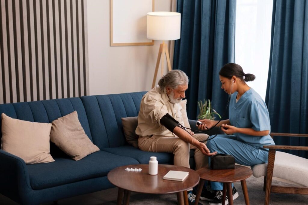 A nurse in blue scrubs measuring the blood pressure of an elderly man sitting on a sofa in a home care setting.