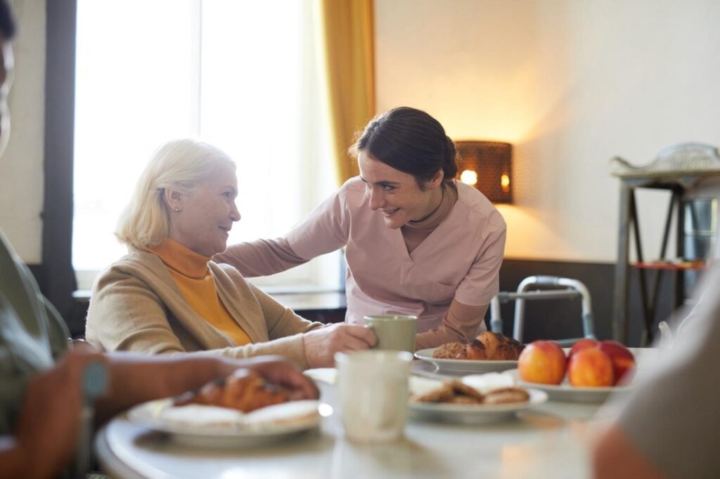 A smiling female caregiver engaging warmly with an elderly woman during breakfast in a communal dining area.