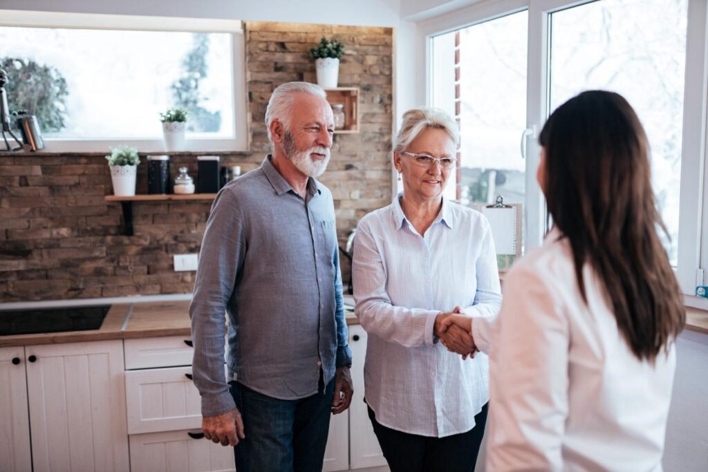 An elderly couple welcoming a female professional into their home, shaking hands in a bright modern kitchen.
