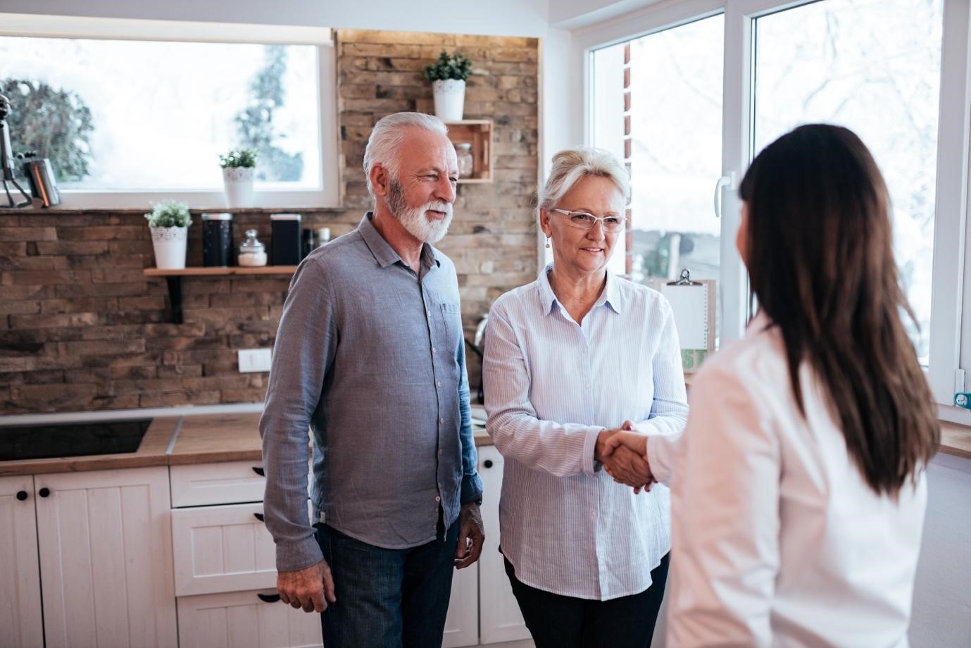 An elderly couple welcoming a female professional into their home, shaking hands in a bright modern kitchen.